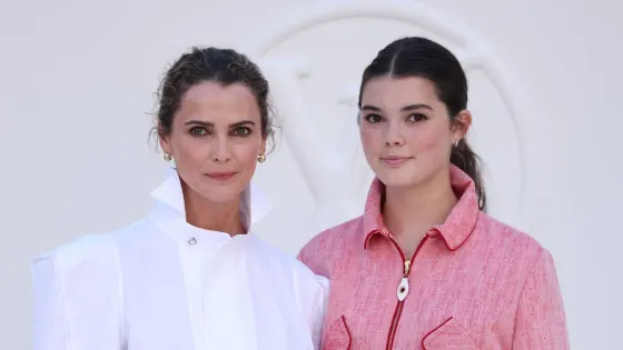 A woman with brunette hair in a tousled bun wearing a white high-neck blouse, black miniskirt and black heels stands next to a teenage girl with brunette hair in a sleek ponytail wearing a pink skirt suit with black heels and a red purse, both smiling at a fashion week event