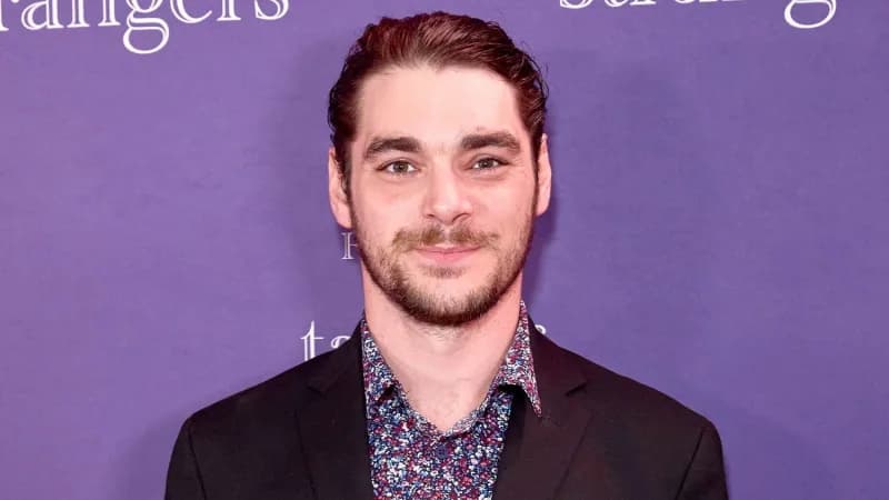 A young man with a warm smile speaking in what appears to be a television interview setting, dressed smartly in a dark jacket