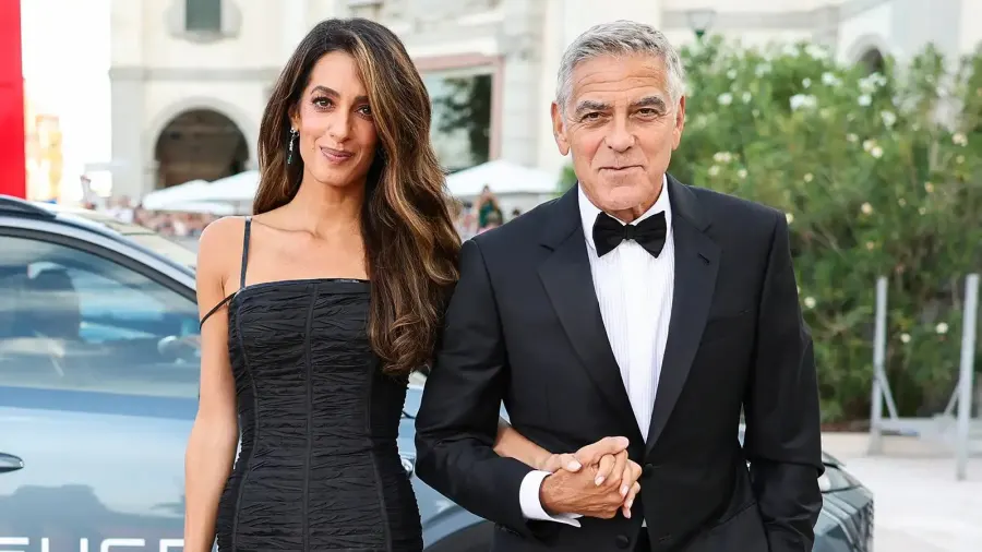 A woman in a sleek black pleated gown with leather details stands on a red carpet at a film festival, her brunette hair styled in soft tousled waves. A man in a dark suit stands beside her.