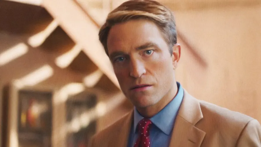 A man in a dark suit stands in what appears to be a television studio setting, looking directly ahead with an intense expression