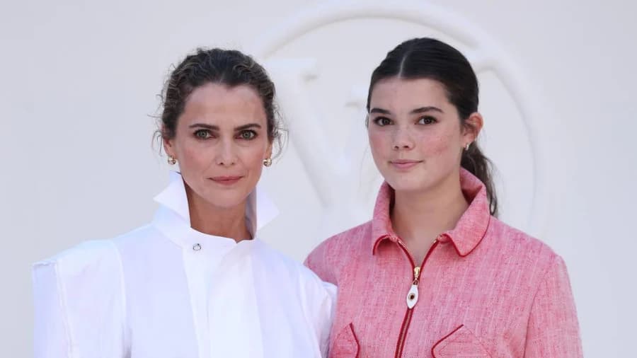 A woman with brunette hair in a tousled bun wearing a white high-neck blouse, black miniskirt and black heels stands next to a teenage girl with brunette hair in a sleek ponytail wearing a pink skirt suit with black heels and a red purse, both smiling at a fashion week event